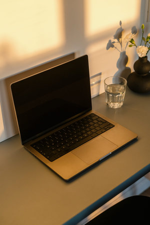 Open laptop on desk in modern room with morning sunlight. High angle view of vase with flowers and...