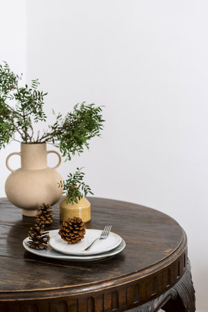 Selective focus on white plates with pine cones on wooden black table top with ceramic vases and green branches. Home decor on christmas. Retro style furniture in apartment. Copy space.の写真素材