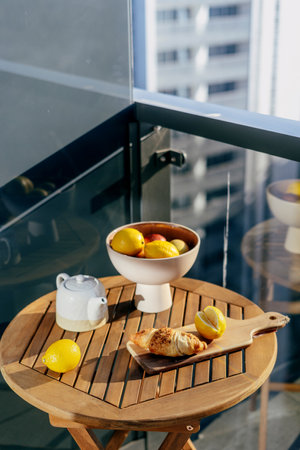 Concepts of slow living. Enjoy morning on balcony. Selective focus on round table with fresh pastry, hot beverage in teapot and ceramic bowl with fruits against cityscape view from private terraceの写真素材