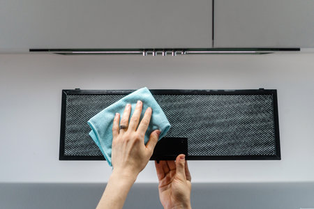 Woman removing black metal air filter from cooker hood for cleaning. Cropped shot of female wiping dirt and dust from kitchen extractor holding textile cloth in handの写真素材