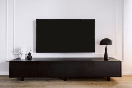 Modern tv screen hanging on white wall in living room with minimalistic interior. Black wooden stand under device. Home decor and lamp on cabinet. Household electronics conceptの写真素材