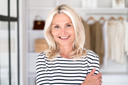 Portrait of smiling middle age woman fashion designer standing against new clothes collection on rack. Happy mature female stylist in front of open wardrobe at homeの写真素材