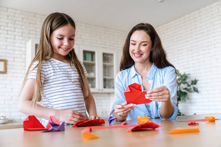 Smiling mother and daughter creating hearts out of red paper in shape of heart, sitting at table in dining room. Free time for hobby activity. Playing with kid at home.の写真素材