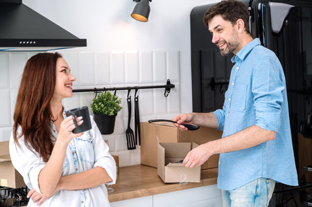 Spouses buy or rent property. Happy smiling couple moving to new house, unpacking boxes with things while standing together at kitchen, drink coffee and talking about furnishing apartmentの写真素材