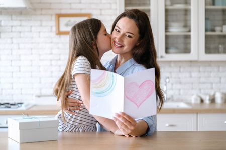 Smiling mother hugging daughter and holding handmade card with heart. Child making surprise to mom, kissing parent, expressing love. Happy mothers day celebration conceptの写真素材