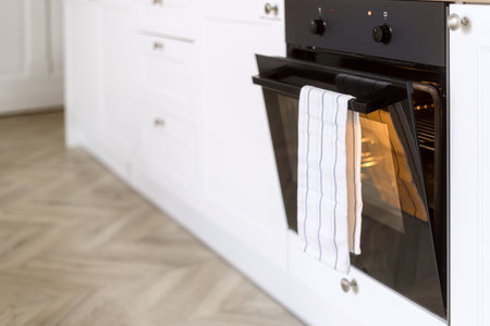 Electric oven with ajar door and light inside, heating up and preparing to cook food. Control panel with temperature mode knob and indicator. Modern household appliance at domestic kitchenの写真素材