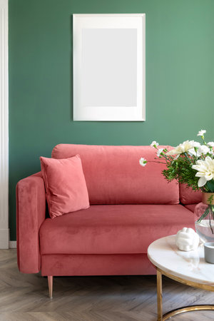Detail in living room interior, comfort couch with decorative pillow, bloom flowers in glass vase on round marble side table and blank picture in frame hanging on green wall.の写真素材