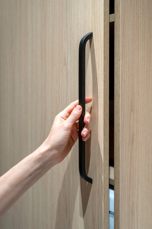 Vertical shot of woman hand closed wooden door of wardrobe or cabinet with shelves. Female holding black handle on furniture. New modern storage system at homeの写真素材