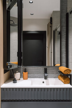 Vertical shot of white ceramic washbasin with black water tap, vanity mirror above sink, clean orange towels folded on shelves in bathroom with modern interior designの写真素材
