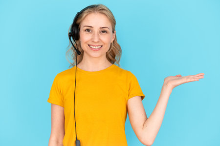 Portrait of helpful woman in headset holding invisible offer on palm, provide client or customer support. Hotline agent worker looking at camera standing isolated on blue copy space backgroundの写真素材