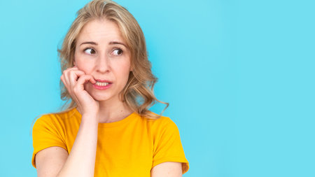 Web banner view of frustrated female feels anxious, bites finger nails in puzzlement, look aside while she standing on blue copy space background. Studio shot of pensive nervous and worried womanの写真素材