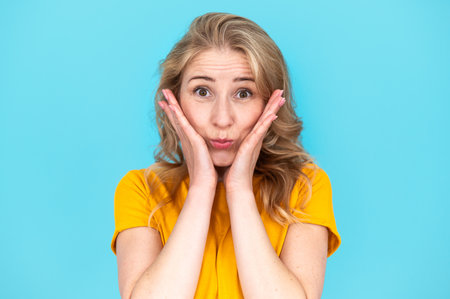 Studio portrait of pretty cute girl looking at camera while she holding her hands close to cheeks and smiling nice. Joyful young woman make silly funny face, standing isolated on blue backgroundの写真素材