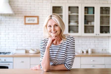 Portrait of mature woman looking at camera and smiling nice. Female sitting at kitchen wooden table against blurred background. Charming housewife in casual clothes spending morning at homeの写真素材