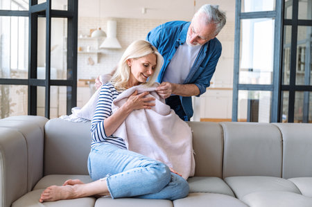 Middle aged man express love and care for lovely wife, giving her warm plaid for comfort rest on couch at home. Mature romantic couple spending time together at cozy living roomの写真素材