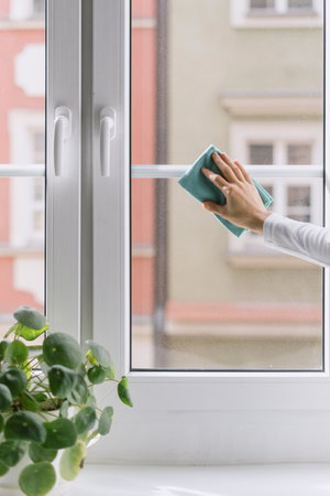 Cropped view of female hand with blue cloth washing glass of closed window with white plastic frame at home. Housewife remove dust and dirty with rag. Cleaning service or advertising concept.の写真素材