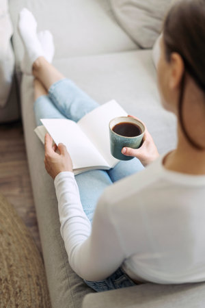 Back rear view of young woman in casual wear sit on comfortable sofa, hold coffee cup in hand, read paper book. Vertical shot of female enjoying her leisure activity resting on couch in living roomの写真素材