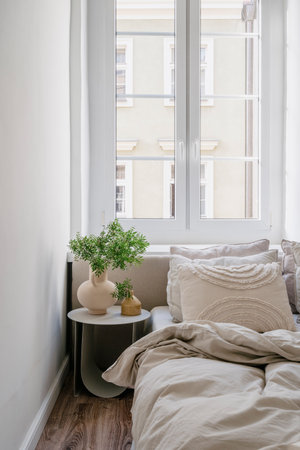 Night table with decorative ceramic vases with green branches next to comfortable bed against window in bedroom in cozy apartment. Bright classic interior design at home.の写真素材