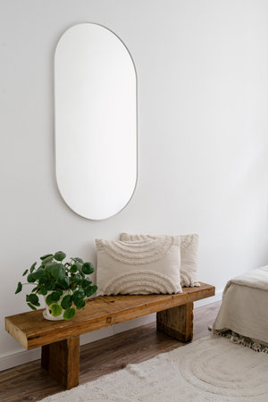 Bedroom space with pilea houseplant in flower pot and cushion on wooden bench under wall mirror. Vertical shot of home decor in minimalist living room interior. Morning at cozy apartment placeの写真素材