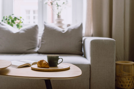 Selective focus on bamboo tray with ceramic cup with hot beverage and fresh bakery on coffee table against blurred living room interior at modern home. Time to rest and read bookの写真素材