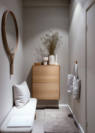 Vertical shot of entrance hall with cushion on comfy bench under mirror, close to shoe cabinet with flowers in vase and home decor. Cozy space, apartment with minimalist interiorの写真素材