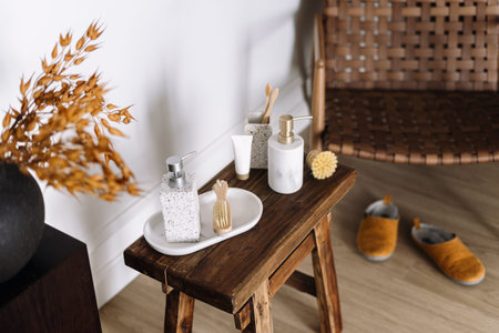 High angle view of soap dispenser bottle on marble tray, toothbrushes, toothpaste and message brush on small wooden bench. Bohemian bathroom interior with rattan chair, home slippers and decorの写真素材