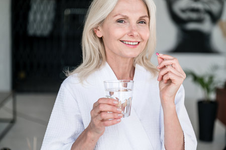 Smiling mature woman holding pill and glass of fresh water on hands and looking at camera. Taking daily medicines, vitamins, supplements. Strengthen skin, hair, nails. Beauty, healthcare conceptの写真素材