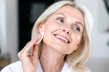 Middle aged woman applying facial hydrating tonic with cotton pad, looking at mirror at bathroom, smile and enjoy skincare routine. Female using anti wrinkle cosmetic productの写真素材