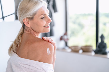 Back view on mature woman put anti aging moisturizer cream on shoulder after shower in bathroom. Female doing morning beauty procedures and using hydrating cosmetic. Bodycare concept.の写真素材