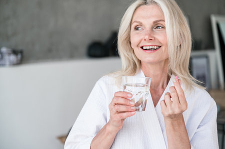 Diet and nutrition concept. Healthy food. Happy smiling 50s old woman taking capsule with Omega 3. Holding glass of water. Vitamin C, D, E, fish oil.の写真素材