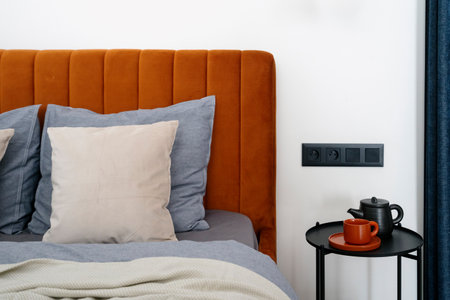 Comfortable bed with soft upholstered headboard, bedding linen, pillows and teapot with ceramic cup on metal side table in stylish hotel room. Bedroom with modern interior and contemporary furniture.の写真素材