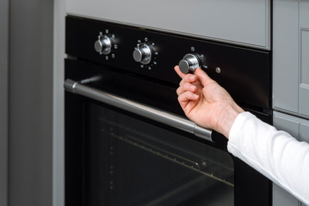 Selective focus on woman hand turning on regulator button of temperature on black new clean electric oven in modern kitchen with built-in equipment at home. Cooking processの写真素材