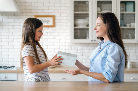 Happy motherhood or custody concept. Smiling daughter making surprise for mother. Woman taking wrapped present gift box from kid in kitchen at home.の写真素材