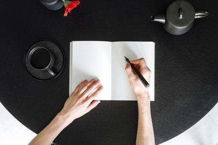 Top view on female hands hold pen and prepare to write in open paper notebook at desk. Woman start note idea in personal diary while sitting at round table with cup of tea and teapot at home.の写真素材