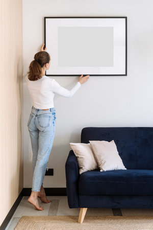 Full length view of woman in casual wear decorated cozy home with art work in picture frame. Copy space poster in living room over comfortable couch. Female install placard on wall, back rear viewの写真素材