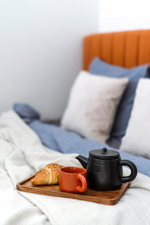 Selective focus on ceramic teapot with brewed herbal tea, cup and fresh french croissant on wooden tray on comfort bed with bedding and pillows at bedroom in morning. Breakfast concept.の写真素材