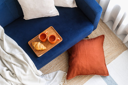 Top view on couch with blue upholstery, soft plaid, decorative pillows, rug on floor and wooden breakfast tray with cups and fresh french croissant in cozy living room. Detail in home interiorの写真素材