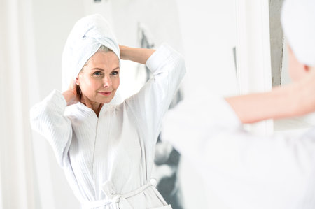 Calm mid age woman in spa bathrobe and towel relaxing after taking shower or bath at home. Reflection in mirror. Beauty, wellness treatment concept. Body and hair care in bathroom.の写真素材
