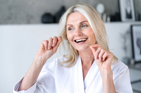 Smiling mature woman in shower robe in bathroom using dental floss for clean teeth and fresh breath. Female doing morning hygiene routine. Oral care conceptの写真素材