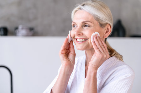 Middle aged smiling woman holding cotton pad disk and cleaning face skin with cleanser. Happy female removing makeup and look away. Satisfied from skincare beauty treatment conceptの写真素材