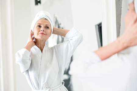 Mature woman standing in front of mirror, wearing a bathrobe and towel after shower, look at reflection, enjoying morning in bathroom. Concept of bodycare, skincare, haircare treatment procedureの写真素材