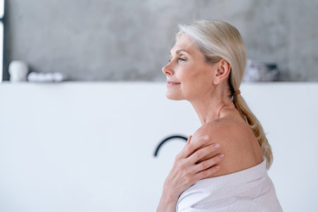 Profile portrait of mature woman in bathrobe standing in white bathroom with copy space on background. Concepts of beauty treatment, home spa, self care, morning beauty routine.の写真素材