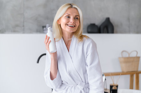Happy middle aged woman in bathrobe hold dispenser bottle with shower gel in bathroom. Smiling female using professional cosmetic product for skin care. Beauty treatment conceptの写真素材