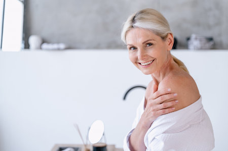 Smiling happy fifty years old woman in robe enjoyed after spa salon body care procedures. Soft mature skin after moisturizer lotion, cream or balm. Copy space. Wellness concept.の写真素材
