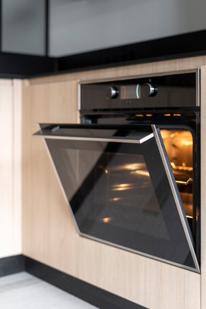 Vertical shoot of open door on new oven with light inside. Black modern household appliance built in wooden cabinet in scandinavian style. Concepts of kitchen design, equipment for cooking baking foodの写真素材