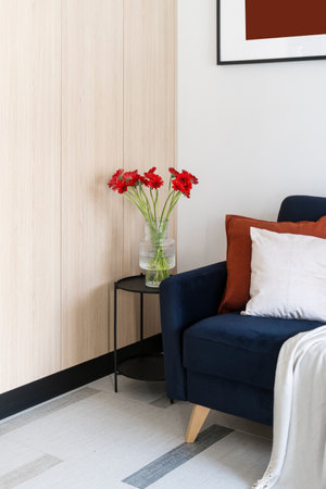 Vertical shot of comfortable blue armchair with pillows and plaid, coffee table with glass vase with flowers next to wooden beige wardrobe in cozy living room. Classic style in home interior.の写真素材