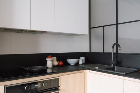 Modern kitchen with built in appliances, electric cooker hood, stove, oven and sink. Frying pan on glass ceramic surface at black marble countertop close to home decor under white cupboardsの写真素材