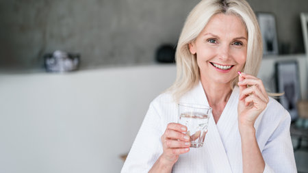 Web banner view of happy mature woman holding glass of still water, and vitamin pill with calcium and collagen. Mid age female feel good, smiling nice, taking dose of daily multivitamins at homeの写真素材