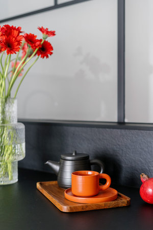 Closeup on orange cup with saucer and ceramic teapot on wooden tray near glass vase with red bloom flowers on black countertop at modern kitchen. Preparing herbal beverage for breakfastの写真素材