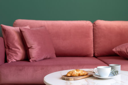 Selective focus on marble countertop of coffee table with cup and wooden board with croissant against red comfortable sofa in modern living room. Breakfast at home.の写真素材