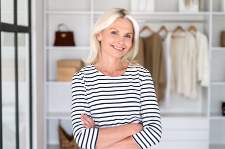 Smiling middle aged woman standing in front of open wardrobe with neatly hanging clothes. Confident female stylist with crossed arms in fashionable boutique present new collection.の写真素材